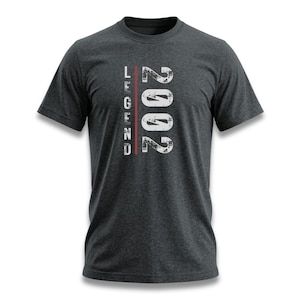 May include: Dark grey t-shirt featuring the text "LEGEND 2002" in a distressed white font. The word "LEGEND" is vertically aligned on the left, with "2002" on the right. A red vertical line separates the two.