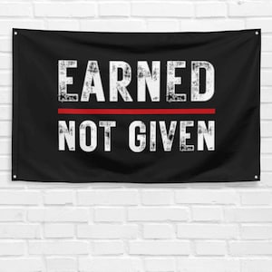 May include: Black flag with the words "EARNED" above a red line and "NOT GIVEN" in distressed white lettering. The flag is hanging against a white brick wall.