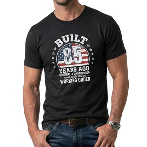 May include: Dark grey t-shirt featuring a distressed graphic with the text "BUILT 85 YEARS AGO" and an American flag design. The shirt also includes the text "ORIGINAL & UNRESTORED most parts still in WORKING ORDER."