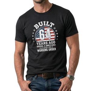 Peut inclure: T-shirt gris foncé avec l'inscription blanche "BUILT 64 YEARS AGO", accompagnée d'un motif rouge, blanc et bleu. Le t-shirt comporte également le texte "ORIGINAL & UNRESTORED most parts still in WORKING ORDER".