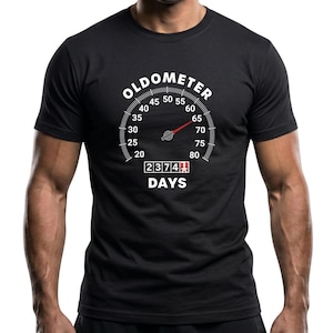 May include: Black t-shirt with a white graphic of a speedometer labeled "OLDOMETER". The gauge reads 65, and the counter below displays "2374 DAYS". The shirt is a crew neck.