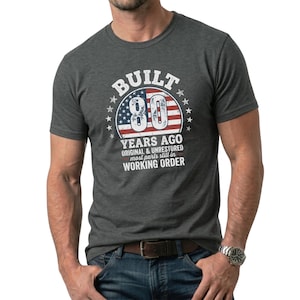 Op de afbeelding: Donkergrijs t-shirt met een afbeelding met de tekst "BUILT 80 YEARS AGO ORIGINAL & UNRESTORED most parts still in WORKING ORDER" in wit en rood, met een sterren- en strepenontwerp. Het t-shirt wordt door een persoon gedragen.
