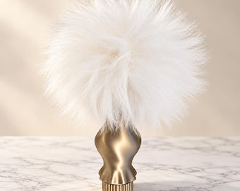 May include: A decorative finial featuring a large, fluffy white faux fur pom-pom attached to a smooth, metallic gold-toned base with a fluted, cylindrical bottom section. The object has a rounded, hourglass-shaped neck and a textured base.