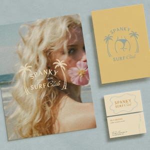 May include: A promotional image featuring stationery for the "Spanky Surf Club." The items include a large poster with a woman's face and the club's logo, a yellow notebook, and a business card. The logo features palm trees and a stylized illustration.
