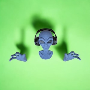 Peut inclure: Une figurine d'extraterrestre grise portant un casque audio noir, les bras tendus sur un fond vert.