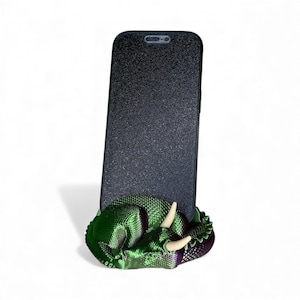 Puede incluir: Un teléfono inteligente negro está sostenido por un soporte para teléfono con forma de dragón verde y morado. El dragón tiene cuernos blancos y una apariencia texturizada y escamosa. El teléfono tiene una superficie negra texturizada.