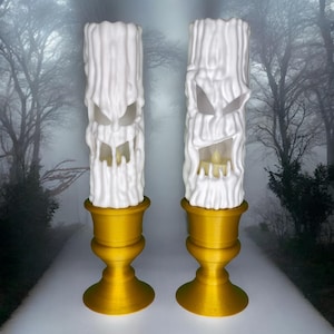 Könnte beinhalten: Zwei weiße, baumstammförmige Kerzenhalter mit geschnitzten Gesichtern, auf goldfarbenen Sockeln. Ein Halter hat eine brennende Kerze im Inneren. Der Hintergrund ist ein verschwommenes Bild eines Waldes, das ein gruseliges Halloween-Thema erzeugt.
