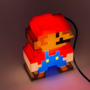 Puede incluir: Una l&aacute;mpara de personaje pixelado con gorra roja, cara color canela y peto azul. La l&aacute;mpara emite un brillo c&aacute;lido desde el interior, resaltando el dise&ntilde;o de bloques de 8 bits. Un cable de alimentaci&oacute;n negro sale del lado de la figura.