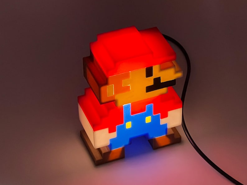 Puede incluir: Una l&aacute;mpara de personaje pixelado con gorra roja, cara color canela y peto azul. La l&aacute;mpara emite un brillo c&aacute;lido desde el interior, resaltando el dise&ntilde;o de bloques de 8 bits. Un cable de alimentaci&oacute;n negro sale del lado de la figura.