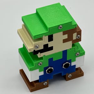 Puede incluir: Una figura pixelada y cuadrada de un personaje con gorra verde, camisa verde, peto azul y zapatos marrones. La figura est&aacute; construida con piezas de pl&aacute;stico superpuestas de varios colores, unidas por tornillos met&aacute;licos plateados visibles.