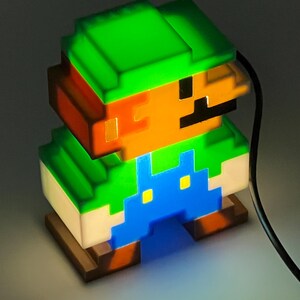 Puede incluir: Una l&aacute;mpara de personaje pixelado con sombrero verde, cabello casta&ntilde;o, bigote negro y peto azul. La figura est&aacute; iluminada desde el interior, emitiendo un brillo intenso a trav&eacute;s de la estructura de pl&aacute;stico transl&uacute;cido, con un cable negro.