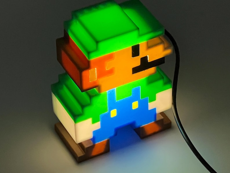 Puede incluir: Una l&aacute;mpara de personaje pixelado con sombrero verde, cabello casta&ntilde;o, bigote negro y peto azul. La figura est&aacute; iluminada desde el interior, emitiendo un brillo intenso a trav&eacute;s de la estructura de pl&aacute;stico transl&uacute;cido, con un cable negro.