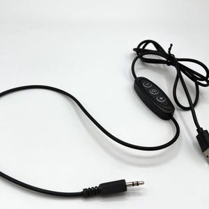 Puede incluir: Un cable de alimentaci&oacute;n USB negro con un m&oacute;dulo de control integrado que tiene tres botones circulares marcados con un signo menos, un s&iacute;mbolo de encendido y un signo m&aacute;s. El cable termina en un conector USB-A y un jack de audio de 3,5 mm.
