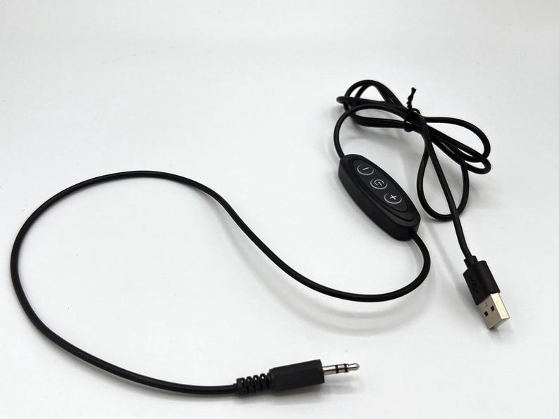 Puede incluir: Un cable de alimentaci&oacute;n USB negro con un m&oacute;dulo de control integrado que tiene tres botones circulares marcados con un signo menos, un s&iacute;mbolo de encendido y un signo m&aacute;s. El cable termina en un conector USB-A y un jack de audio de 3,5 mm.