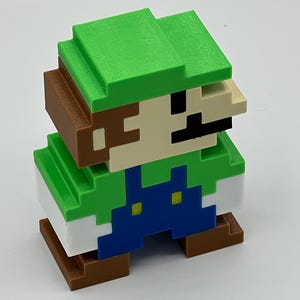 Puede incluir: Una figura 3D pixelada de un personaje con sombrero verde, camisa verde, peto azul con dos botones amarillos y zapatos marrones. La figura tiene la cara de color beige con un bigote negro y pelo casta&ntilde;o, con un estilo c&uacute;bico y bloqueado.