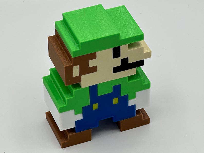 Puede incluir: Una figura 3D pixelada de un personaje con sombrero verde, camisa verde, peto azul con dos botones amarillos y zapatos marrones. La figura tiene la cara de color beige con un bigote negro y pelo casta&ntilde;o, con un estilo c&uacute;bico y bloqueado.