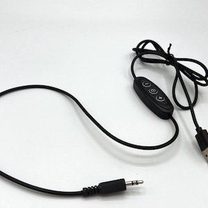 Puede incluir: Un cable de alimentaci&oacute;n USB negro con un m&oacute;dulo de control integrado que tiene tres botones circulares marcados con un signo menos, un s&iacute;mbolo de encendido y un signo m&aacute;s. El cable termina en un conector USB-A y un jack de audio de 3,5 mm.