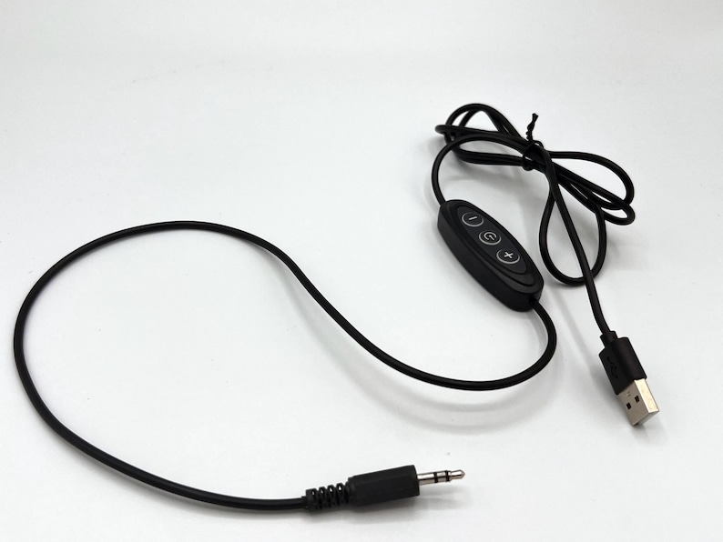 Puede incluir: Un cable de alimentaci&oacute;n USB negro con un m&oacute;dulo de control integrado que tiene tres botones circulares marcados con un signo menos, un s&iacute;mbolo de encendido y un signo m&aacute;s. El cable termina en un conector USB-A y un jack de audio de 3,5 mm.