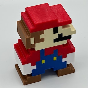 Puede incluir: Una figura pixelada en tres dimensiones de un personaje con gorra roja, camisa roja, peto azul con botones amarillos y zapatos marrones. La figura presenta una cara de color beige con un bigote negro y est&aacute; formada por bloques c&uacute;bicos.