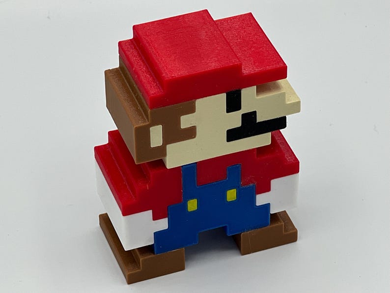 Puede incluir: Una figura pixelada en tres dimensiones de un personaje con gorra roja, camisa roja, peto azul con botones amarillos y zapatos marrones. La figura presenta una cara de color beige con un bigote negro y est&aacute; formada por bloques c&uacute;bicos.