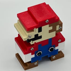 Puede incluir: Una figura 3D pixelada construida con bloques de pl&aacute;stico rojo, azul, blanco, beige y marr&oacute;n. El personaje presenta un sombrero rojo, cara beige con rasgos faciales negros, peto azul y zapatos marrones, unidos por tornillos met&aacute;licos plateados visibles.