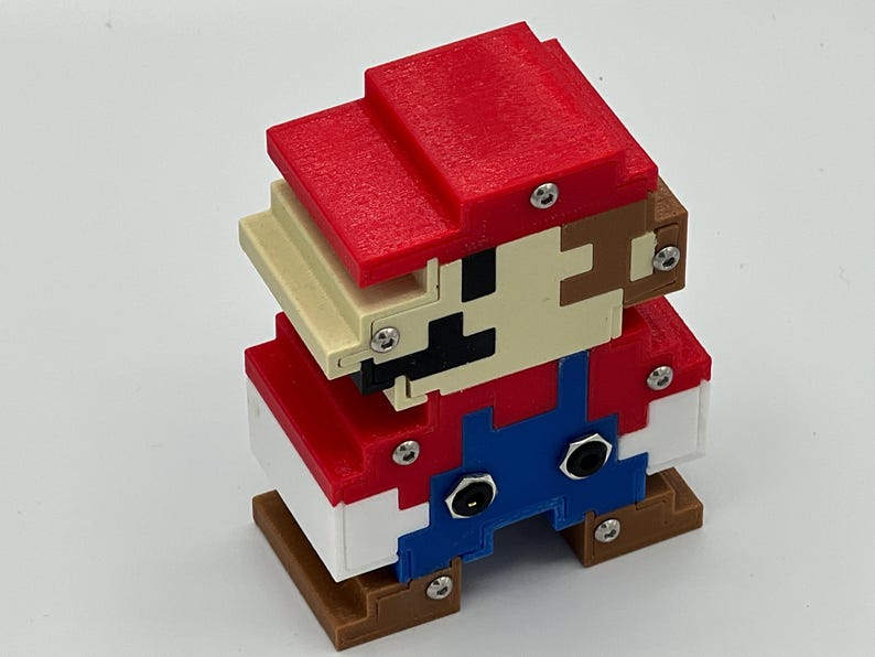 Puede incluir: Una figura 3D pixelada construida con bloques de pl&aacute;stico rojo, azul, blanco, beige y marr&oacute;n. El personaje presenta un sombrero rojo, cara beige con rasgos faciales negros, peto azul y zapatos marrones, unidos por tornillos met&aacute;licos plateados visibles.