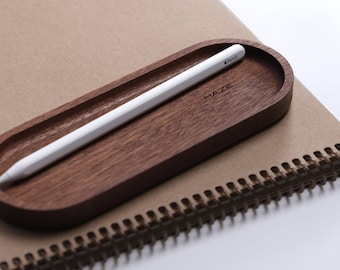 Könnte beinhalten: Ein hölzerner Stifthalter mit einem weißen Apple Pencil darin. Der Halter ist oval geformt und hat die Aufschrift "HAZE" eingraviert.