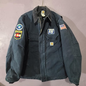 May include: A dark navy blue Carhartt work jacket featuring a corduroy collar, a front zipper, and multiple patches. The right sleeve displays a circular BJ logo and a Colorado state flag patch. The left chest has a BJ logo and a US flag patch.
