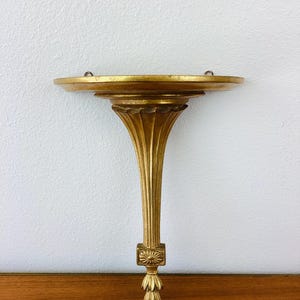 May include: A gold-colored wall shelf with a round top and a decorative, fluted, and ornate base.