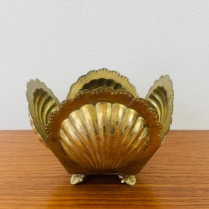 May include: A vintage brass seashell-shaped bowl with a scalloped rim and three small feet. The bowl has a textured surface and a golden hue, suggesting it is made of brass. The design is reminiscent of a clam shell.
