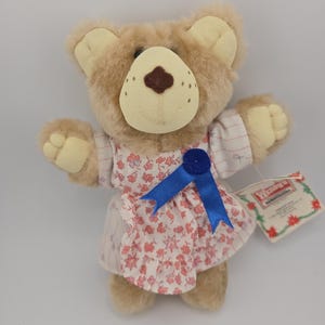 May include: A plush brown teddy bear wearing a pink and white floral dress with a blue ribbon. The bear has a brown nose and yellow paws. The bear is holding a tag that says "Merry's  Manufactures".