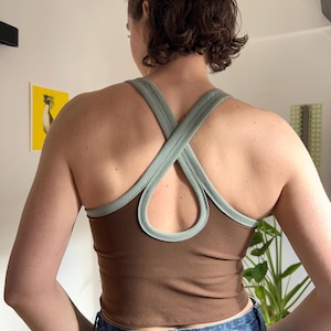May include: A brown tank top with light green trim and cross-back straps. The top is worn with blue denim jeans. The design includes a keyhole back detail, providing a contemporary and stylish appearance.