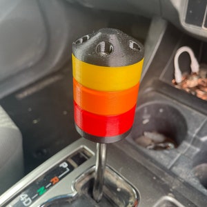 May include: A multi-coloured shift knob with a black top, yellow, orange, and red bands. The knob is cylindrical and sits atop a metal shift lever in a vehicle's interior.