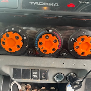 May include: A close-up of a car's dashboard with a black and orange knob for the temperature control, a black and orange knob for the fan speed, and a black and orange knob for the air conditioning. The dashboard also has a USB and AUX port, a cigarette lighter, and a security button.