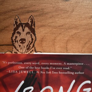 May include: A wooden bookmark featuring a laser-engraved image of a dog's head. The bookmark is placed on a book with the text "It's perfection, every word, every moment. A masterpiece... One of the best books I've ever read." -LISA JEWELL, #1 New York Times bestselling author"