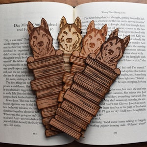 May include: Four wooden bookmarks featuring a dog peeking over a stack of books. The bookmarks are made of light brown wood with a detailed engraved design.