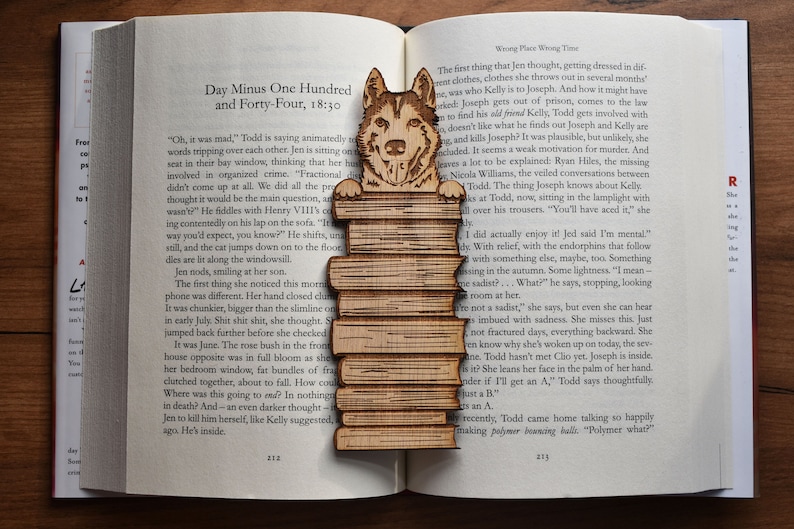 May include: A wooden bookmark featuring a cartoon husky dog peeking over a stack of books. The bookmark is a light brown color and has a detailed design.