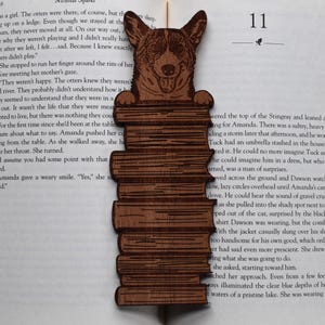 May include: A wooden bookmark featuring a smiling dog's head above a stack of books. The bookmark is made of brown wood and is placed on an open book. The page number "11" is visible.