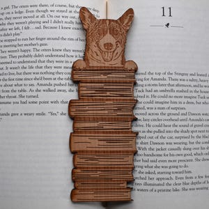 May include: Wooden bookmark featuring a smiling corgi dog design atop a stack of books. The bookmark is made of light brown wood and is placed on the pages of a book, with the page number 11 visible.