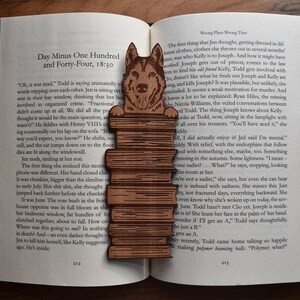 May include: A wooden bookmark featuring a brown and white wolf illustration. The bookmark is layered with multiple wooden planks, creating a textured look. The bookmark is placed on an open book with text visible.