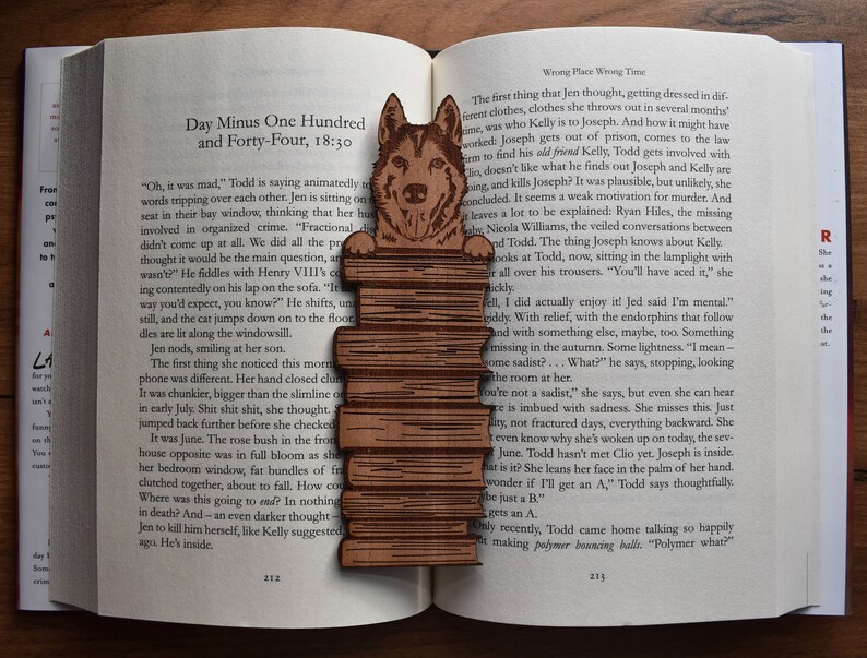 May include: A wooden bookmark featuring a brown and white wolf illustration. The bookmark is layered with multiple wooden planks, creating a textured look. The bookmark is placed on an open book with text visible.
