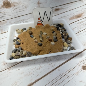 May include: A white rectangular tray containing tan-coloured material, small pebbles, and larger rocks. A small sign with a white background, the letter "W", and an orange traffic cone graphic is placed in the tan material.