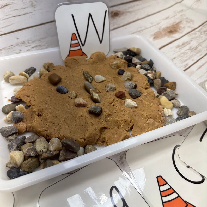 May include: A white tray containing brown playdough and small rocks. A card with the letter "W" and a traffic cone illustration is placed in the playdough. Educational toy for children.