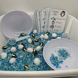 May include: A white plastic tub filled with blue and white rice, white cotton balls, and small penguin figurines. There are also clear plastic ice cubes and a white plastic scoop. A set of cards with the title "Penguin Play" and a list of items to count, such as "9 Scoops of Snow", "4 Penguins", "5 Icebergs", and "6 Eggs".