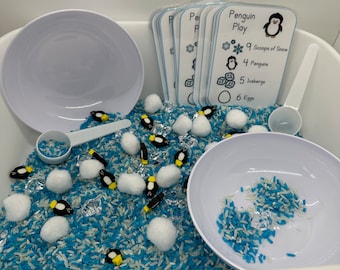 May include: A white plastic tub filled with blue and white rice, white cotton balls, and small penguin figurines. There are also clear plastic ice cubes and a white plastic scoop. A set of cards with the title "Penguin Play" and a list of items to count, such as "9 Scoops of Snow", "4 Penguins", "5 Icebergs", and "6 Eggs".