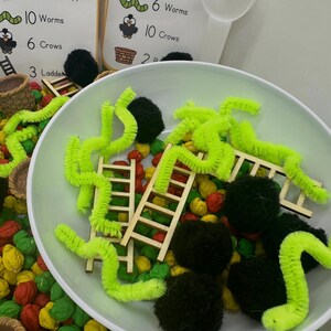 May include: A sensory play setup featuring a white bowl filled with colorful beads, wooden ladders, green pipe cleaner worms, and black pom-poms. Two cards display text: "10 Worms, 6 Crows, 3 Ladders" and "6 Worms, 10 Crows, 2 P".