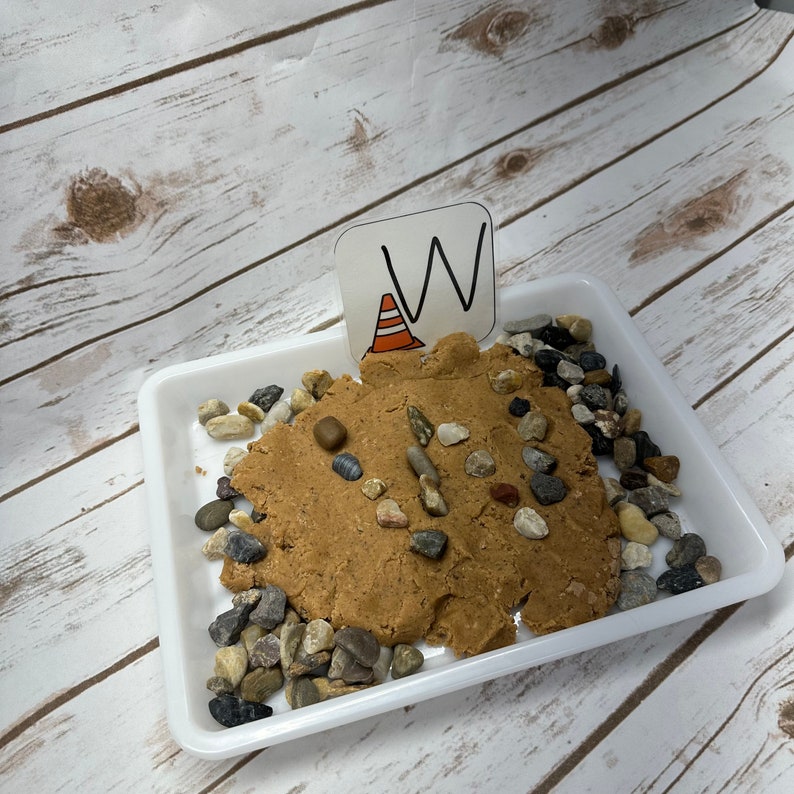 May include: A white plastic tray holds a brown, textured material, dotted with small stones. A small sign with the letter "W" and a traffic cone graphic is placed in the material. The background is a light wood grain pattern.