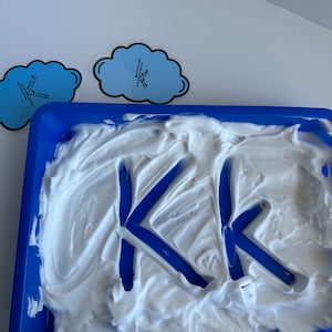 May include: A blue plastic tray filled with white shaving cream. Two blue letters "K" are drawn in the cream.