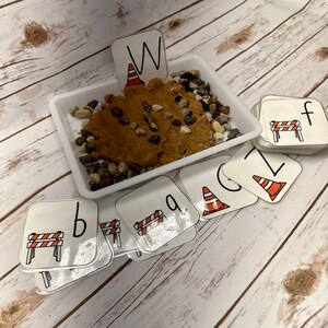 May include: A white tray containing brown playdough, small stones, and letter cards. The cards display letters and construction-themed illustrations, including a traffic cone and a road barrier. The letter "W" is displayed above the playdough.