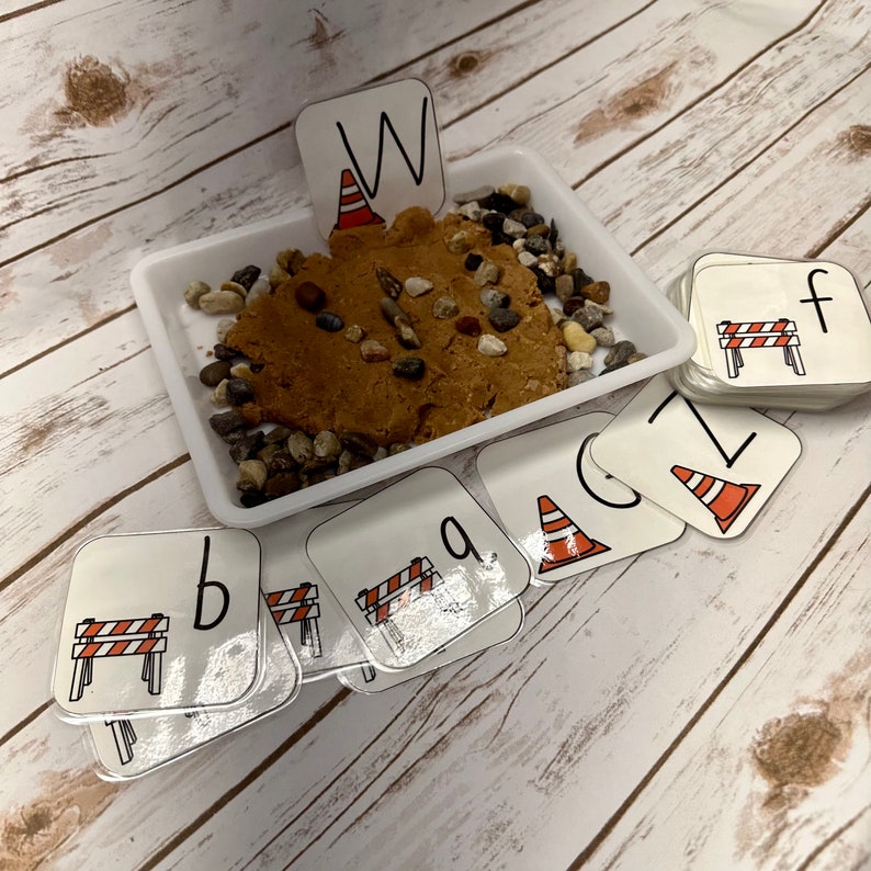 May include: A white tray containing brown playdough, small stones, and letter cards. The cards display letters and construction-themed illustrations, including a traffic cone and a road barrier. The letter "W" is displayed above the playdough.